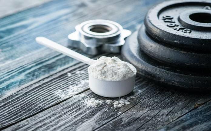 Messlöffel mit Kreatin Pulver neben Hantelscheiben auf Holztisch, Fitness Supplement