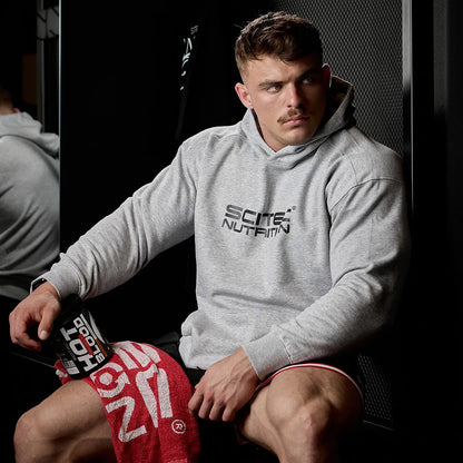 Sportler im Gym mit Scitec Nutrition Hoodie, hält Hot Blood Pre-Workout, Handtuch auf Knie