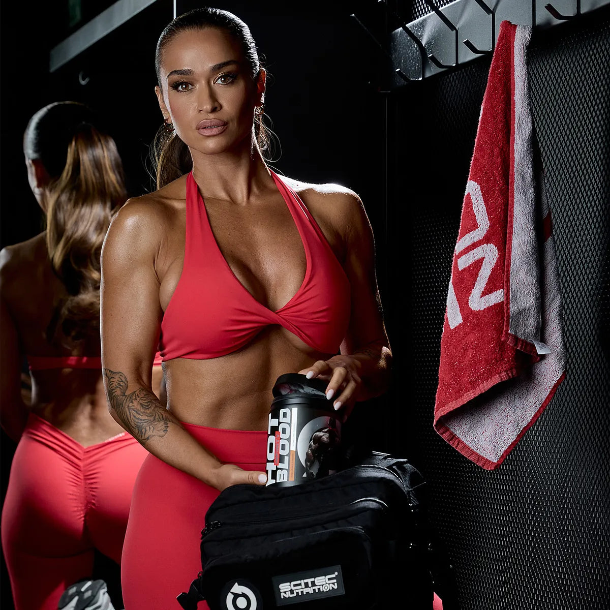Sportliche Frau in rotem Outfit mit Scitec Nutrition Shaker und Sporttasche im Fitnessstudio