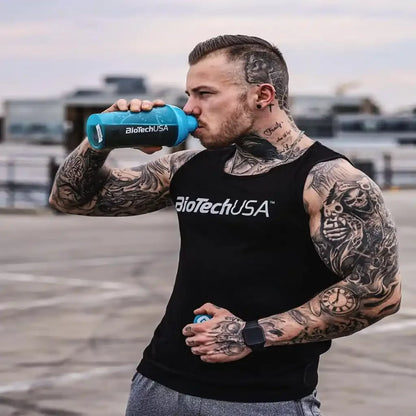 Tattooierter Mann im BioTechUSA Tanktop trinkt aus blauem Shaker auf Parkplatz