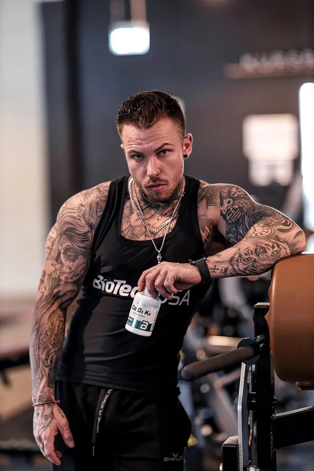 Tattooierter Mann im Fitnessstudio mit BiotechUSA Ca D3 K2 Supplement-Dose