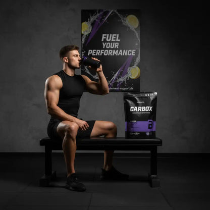 Muskulöser Mann trinkt Fitness-Booster, neben ihm CARBOX-Kohlenhydrat-Pulver für Sportleistung