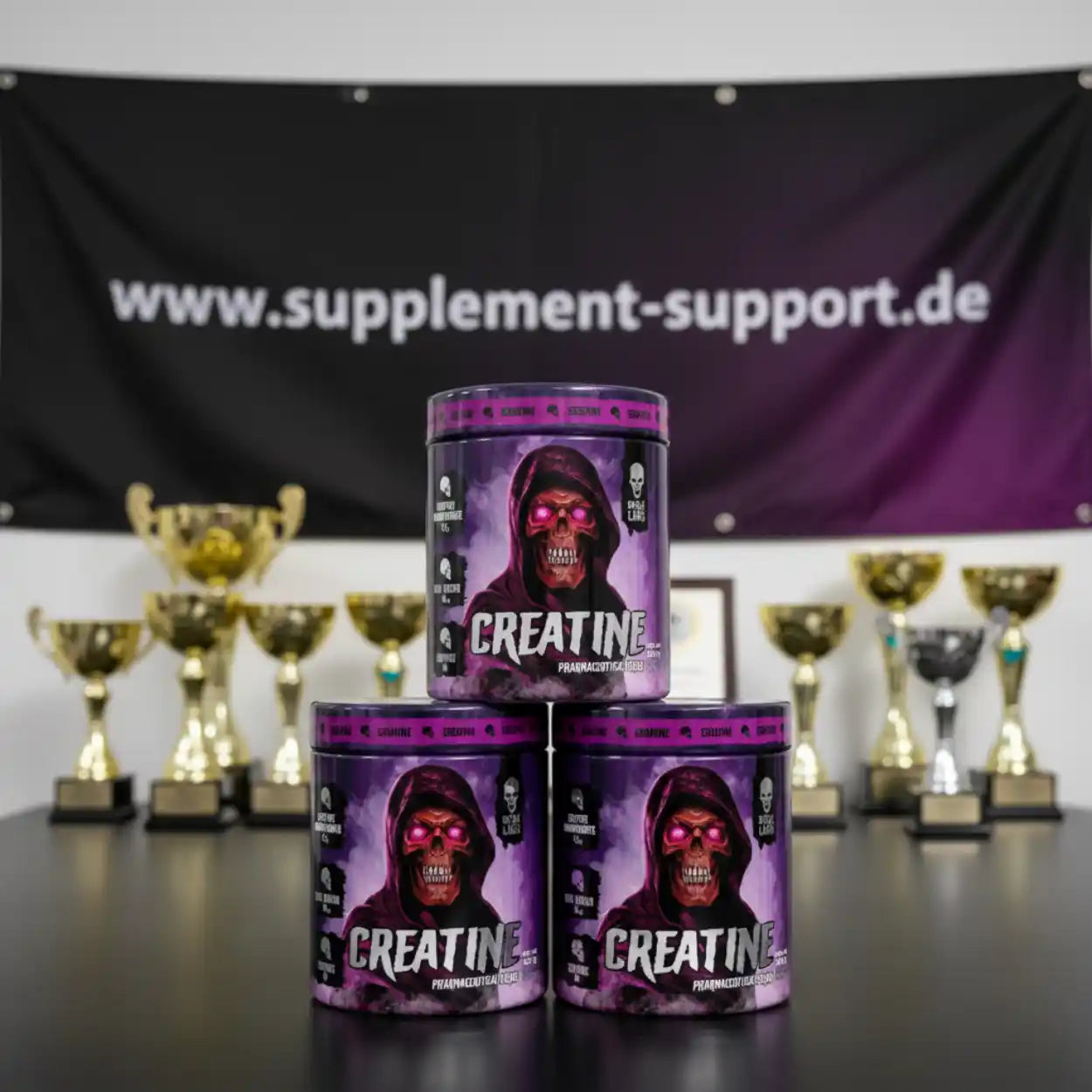 Drei Dosen Kreatin Pulver mit auffälligem Totenkopf-Design vor Pokalen und Banner