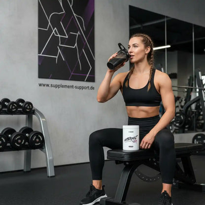 Sportlerin im Fitnessstudio trinkt Shake, Eiweißpulver und Hanteln im Hintergrund
