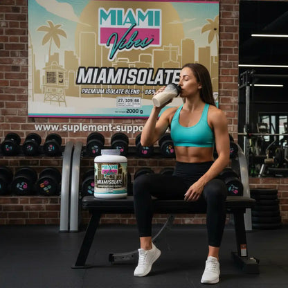 Muskulöse Frau trinkt Proteinshake neben einer Packung MIAMI VIBES MIAMISOLATE im Fitnessstudio.