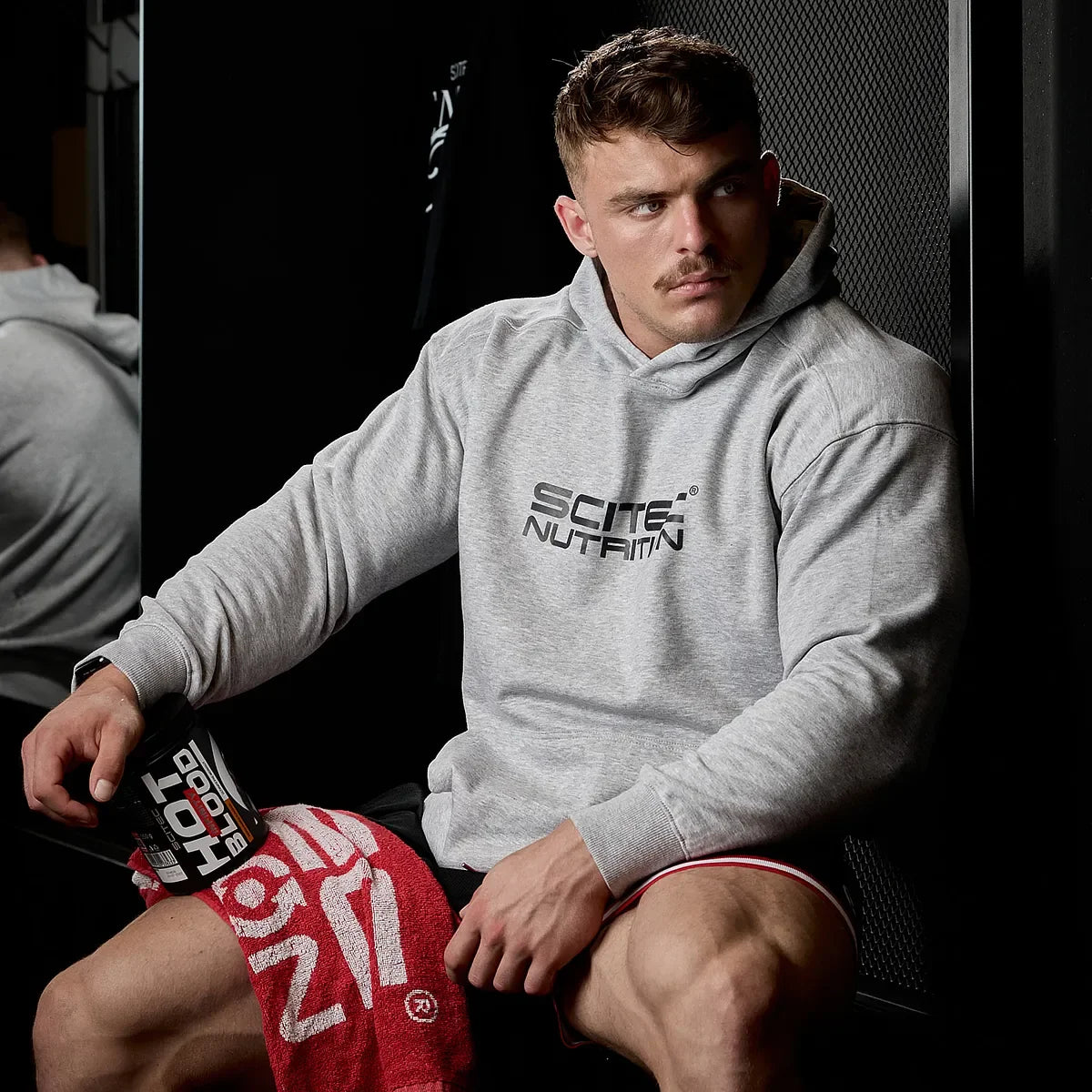 Muskulöser Mann mit Scitec Nutrition Hoodie und Pre-Workout Booster im Fitnessstudio